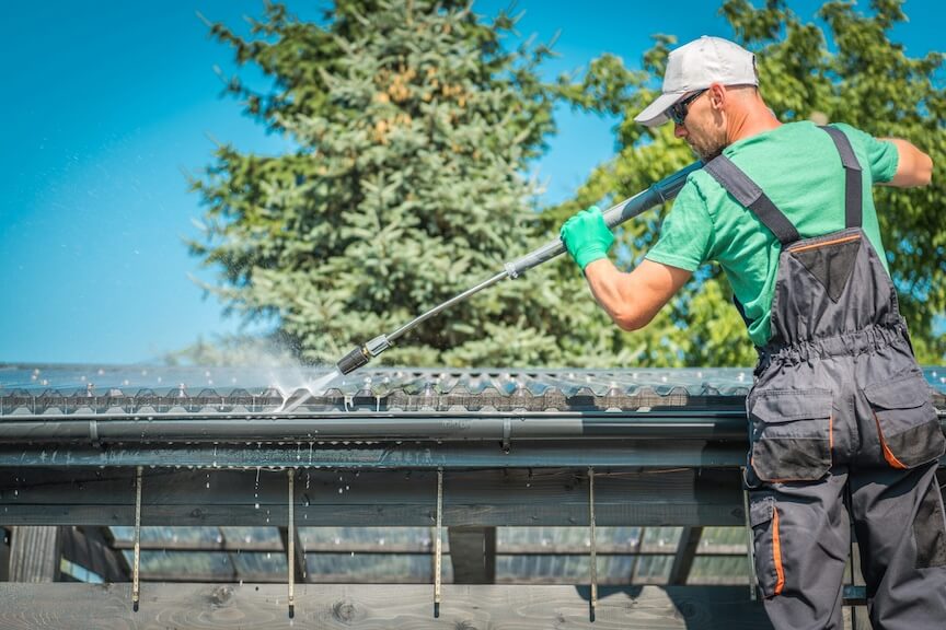 A professional on a ladder using a scoop to clean debris from a gutter